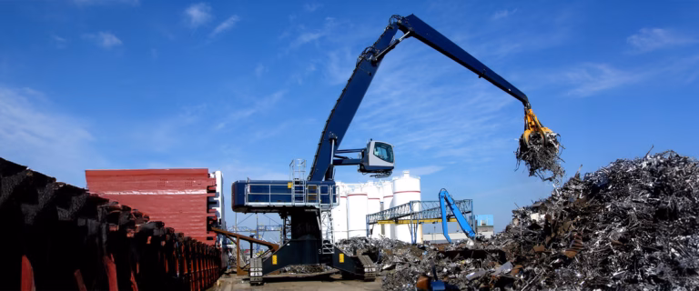 Large blue material handler with grapple arm sorting scrap metal at an industrial yard