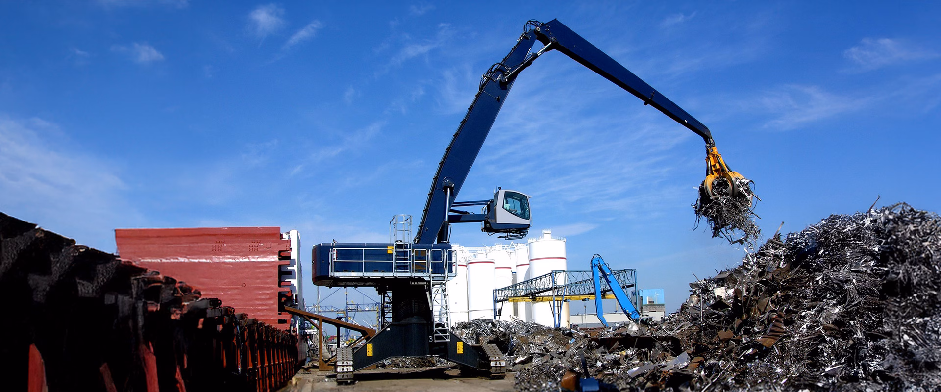 Large blue material handler with grapple arm sorting scrap metal at an industrial yard