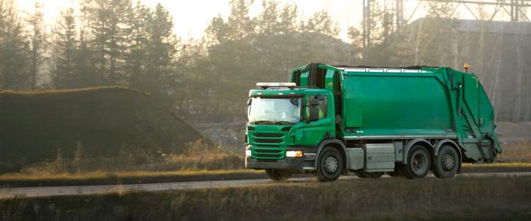 Green Scania refuse collection truck driving on a road near an industrial site