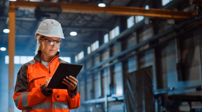 Woman in an orange work jacket, white helmet and safety glasses using a tablet in an industrial hall