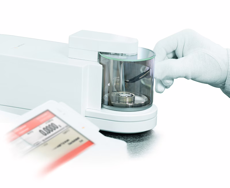 Gloved hand placing a small sample on a laboratory scale inside a glass enclosure