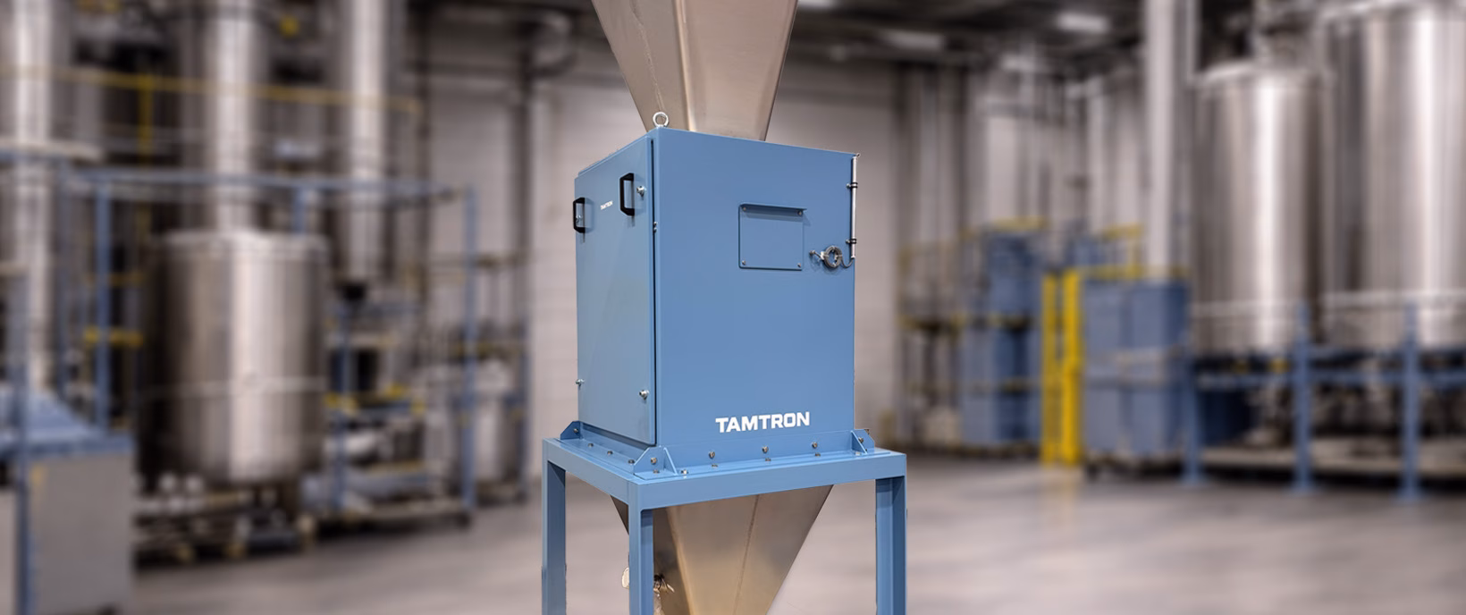 Tamtron totalising hopper scale with blue steel weighing unit and metal cones above and below, against a blurred industrial hall background.