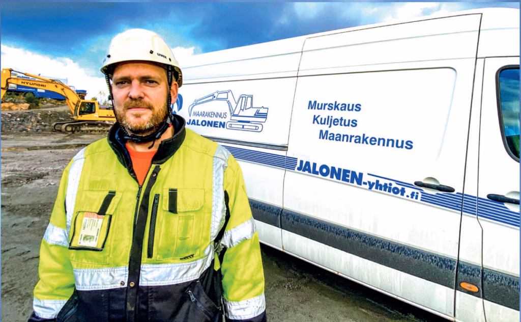 Mies, jolla on valkoinen kypärä ja keltainen työtakki, seisoo työmaalla valkoisen pakettiauton edessä
