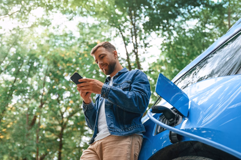 Man using a smartphone while leaning on a charging electric car surrounded by forest scenery