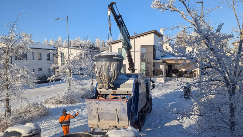 Waste collection truck with a loader crane lifting a large underground waste container in a snowy Finnish residential area on a clear winter day