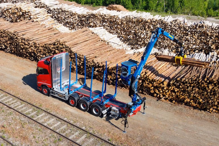 Aerial view of a red timber truck with blue loader crane loading logs at a timber yard